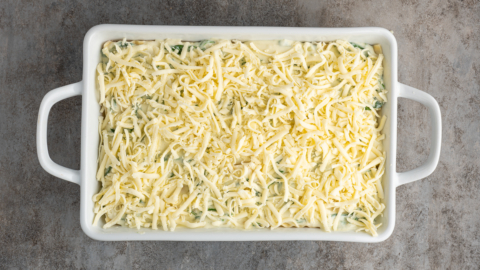 A rectangular white baking dish filled with an unbaked white chicken lasagna recipe, topped with a generous layer of shredded cheese, sits on a gray textured surface.