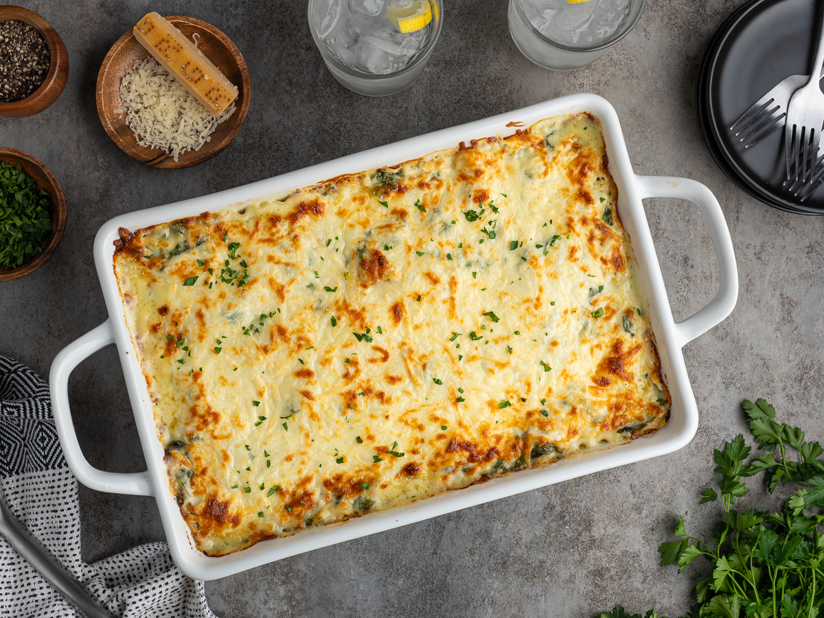 A baked casserole from a chicken lasagna recipe, topped with melted, golden cheese, sits in a white rectangular dish surrounded by grated cheese, chopped herbs, two glasses of iced water with lemon, and utensils on a gray surface.
