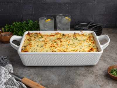 A large white baking dish filled with creamy chicken lasagna, topped with melted cheese and herbs, sits on a gray countertop. In the background are two glasses of water with lemon slices, parsley, and black plates with utensils.