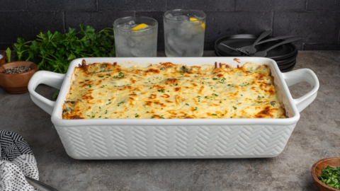 A large white baking dish filled with creamy chicken lasagna, topped with melted cheese and herbs, sits on a gray countertop. In the background are two glasses of water with lemon slices, parsley, and black plates with utensils.