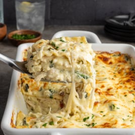 A spatula lifts a cheesy serving of white lasagna from a baking dish, revealing layers of pasta, creamy sauce, spinach, and melted cheese—perfect inspiration for your next chicken lasagna recipe. The dish sits on a gray countertop with bowls and plates nearby.