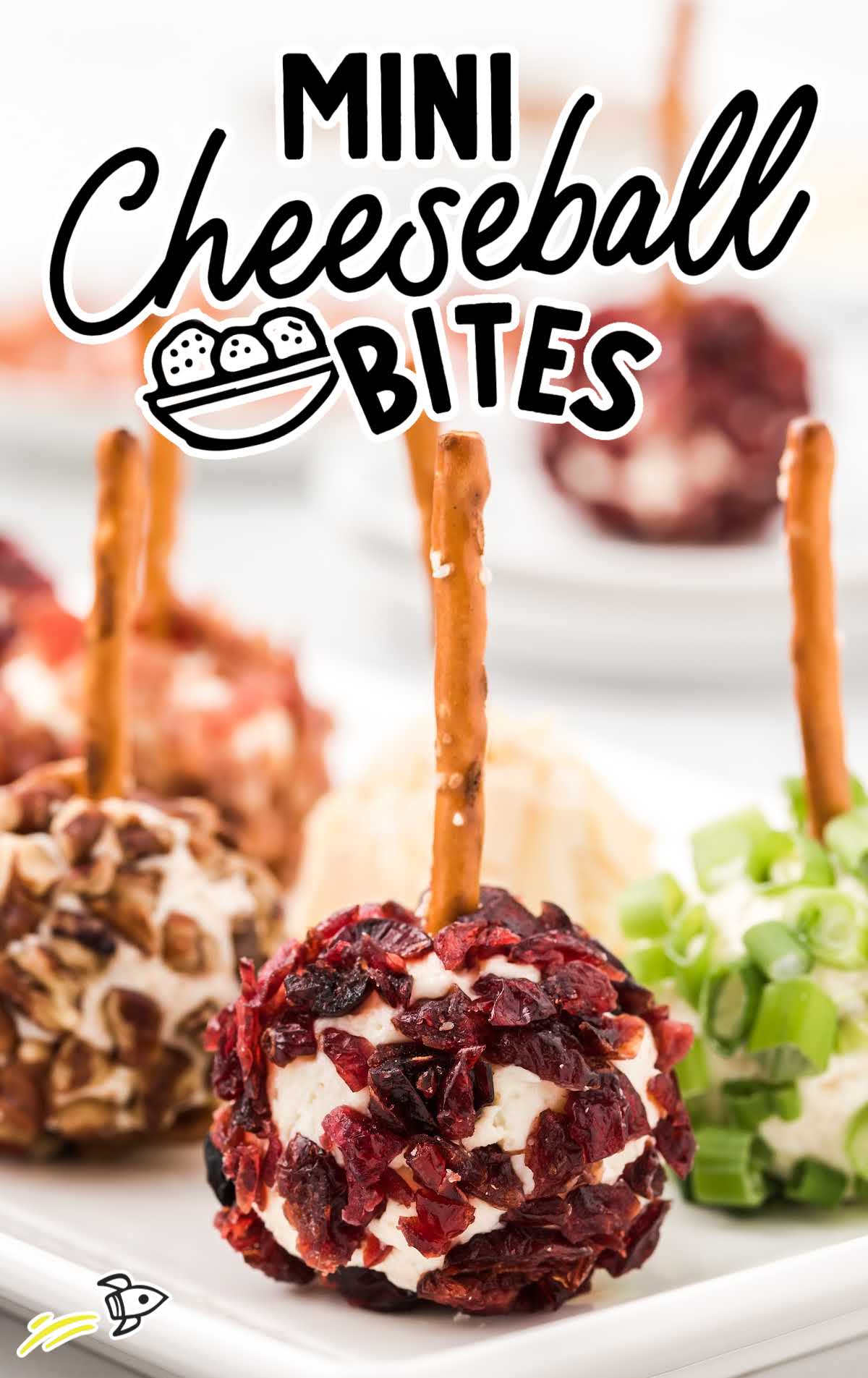 Close-up of mini cheeseball bites with various toppings like cranberries, green onions, and pecans, each with a pretzel stick in the center.