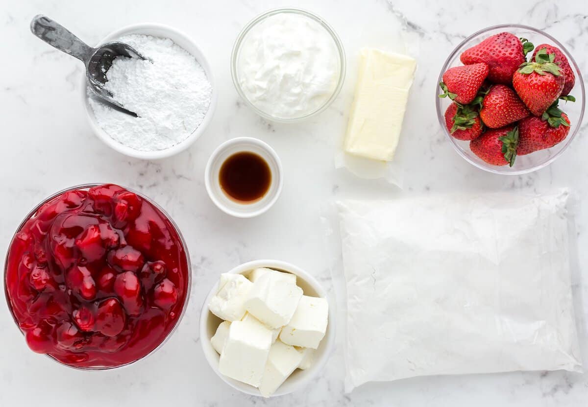 Ingredients on a white countertop for this Strawberry Dump Cake Recipe: powdered sugar, whipped cream, butter, strawberries, cherry pie filling, vanilla extract, cream cheese, and a bag of flour.