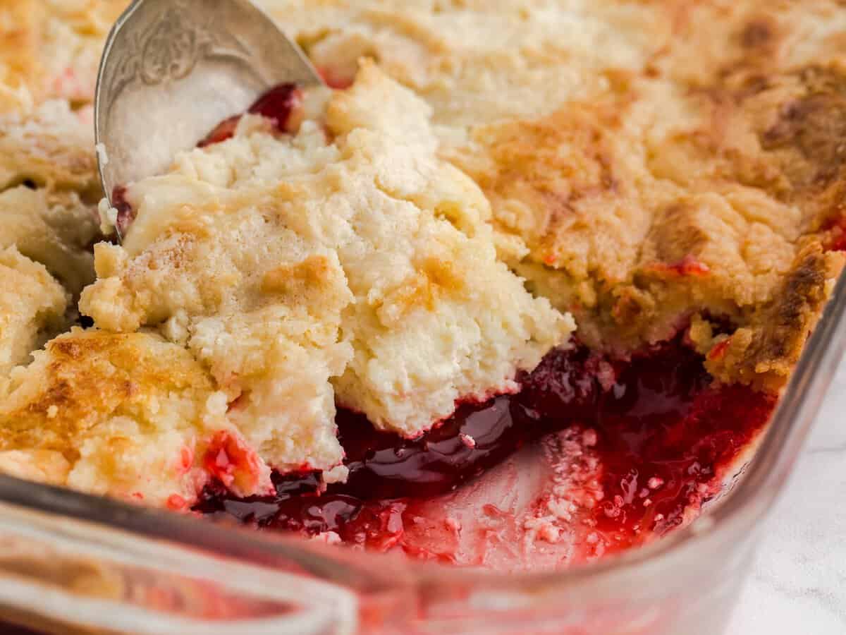 A close-up of a strawberry dump cake in a glass baking dish, with a spoon lifting a portion to reveal the gooey red fruit filling beneath a golden, crumbly crust—an irresistible strawberry dump cake recipe come to life.