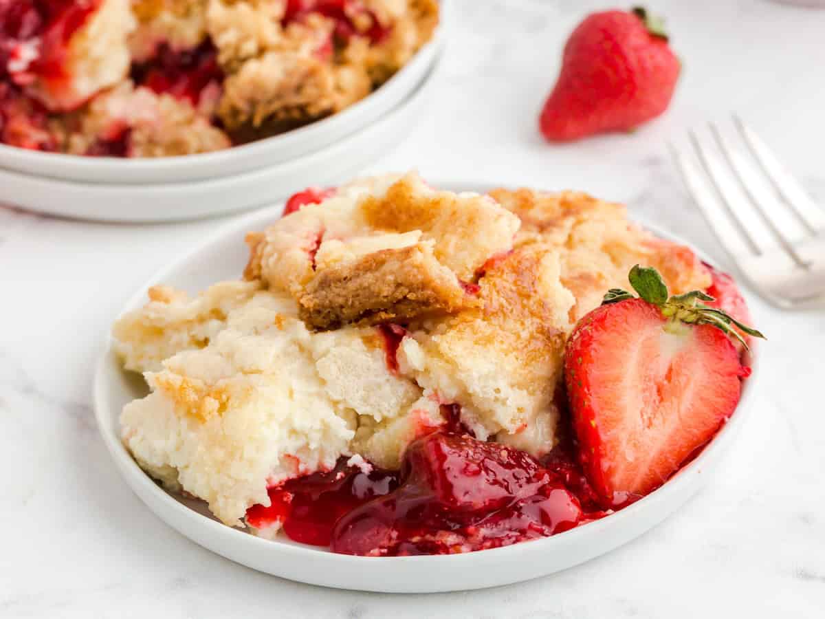 A serving of strawberry cobbler with golden crust and juicy strawberry filling, reminiscent of a homemade Strawberry Dump Cake, garnished with a halved fresh strawberry on a white plate.
