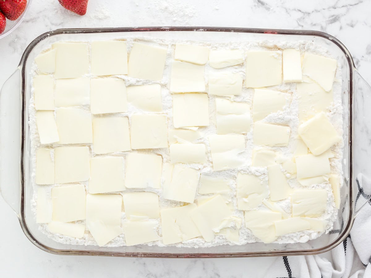 A glass baking dish filled with a white powdery layer, topped evenly with overlapping slices of butter, hints at the start of a delicious Strawberry Dump Cake Recipe; strawberries and a striped towel are nearby.