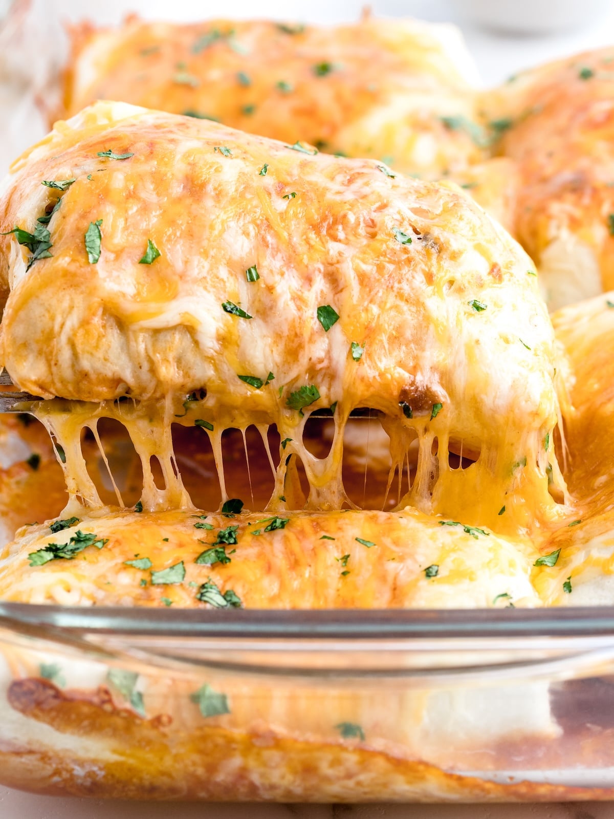 Baked burrito being lifted from a glass casserole dish with a dramatic melted cheese pull