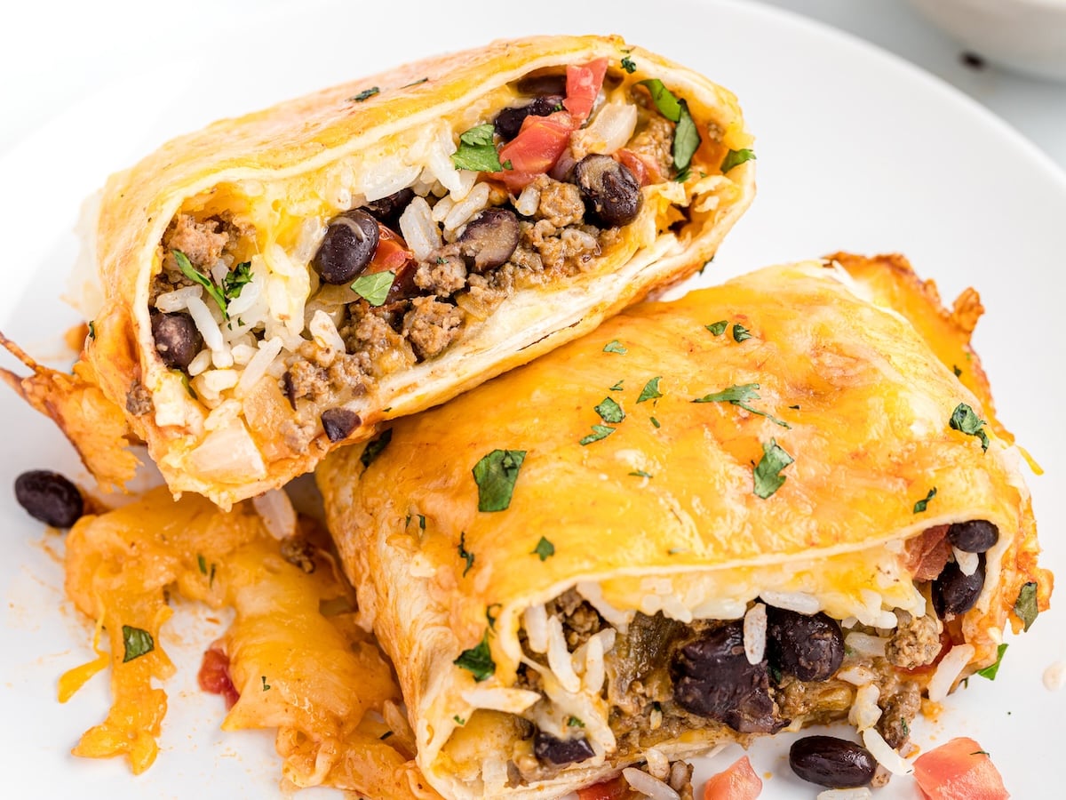 Baked burritos stacked on a white plate, cut open to show a filling of ground beef, black beans, rice, and melted cheese