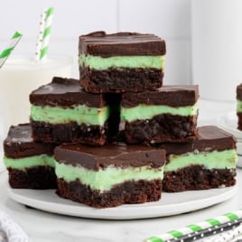 A plate of Mint Chocolate Brownies with three layers: a fudgy brownie base, green mint cream middle, and glossy chocolate top. Striped straws and a glass of milk hint at enjoying this Mint Chocolate Brownies Recipe to the fullest.