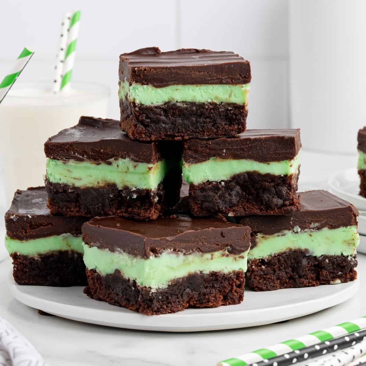A plate of Mint Chocolate Brownies with three layers: a fudgy brownie base, green mint cream middle, and glossy chocolate top. Striped straws and a glass of milk hint at enjoying this Mint Chocolate Brownies Recipe to the fullest.