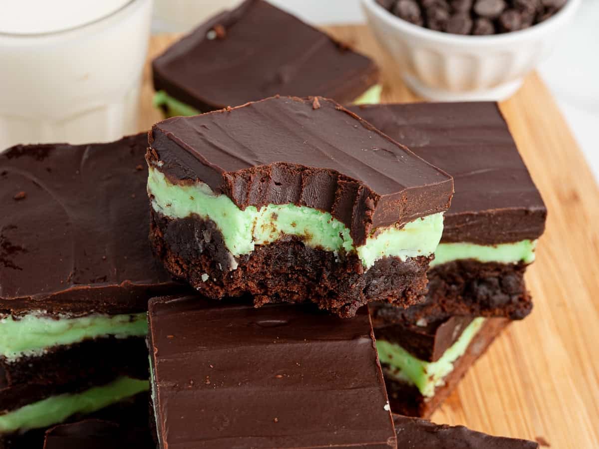A close-up of Mint Chocolate Brownies with layers of dark chocolate, green mint cream, and brownie baseโone with a bite taken outโstacked on a wooden surface next to milk and a bowl of chocolate chips. Try this Mint Chocolate Brownies Recipe!.