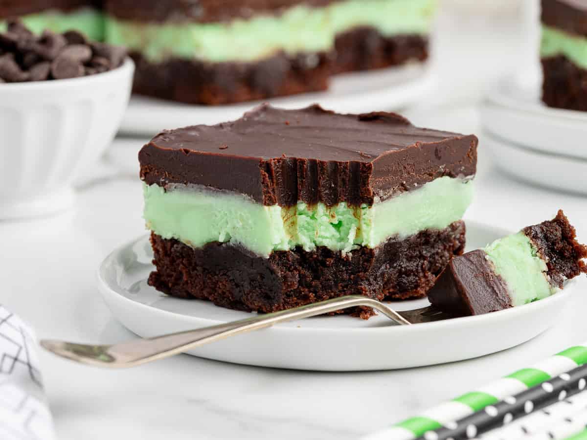 A close-up of a Mint Chocolate Brownie with a bite taken out, showing layers of chocolate ganache, mint cream, and brownie, served on a white plate with a fork and a bowl of chocolate chipsโperfect for any Mint Chocolate Brownies Recipe.