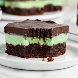 A close-up of a Mint Chocolate Brownies dessert bar, featuring a brownie base, green mint layer, and thick chocolate topping on a white plate with a bite taken out. More treats and a bowl of chocolate complete this Mint Chocolate Brownies Recipe scene.