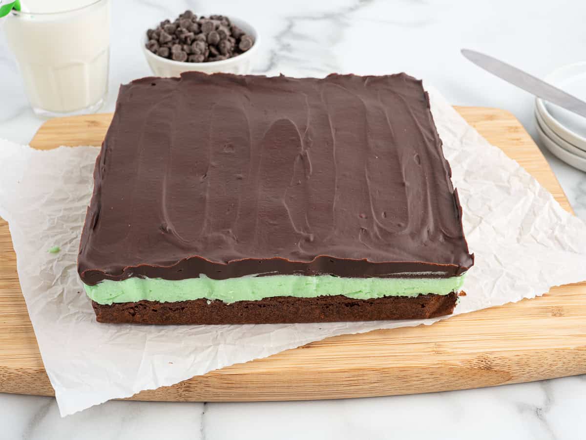 A square Mint Chocolate Brownie with layers of brownie, green mint filling, and smooth chocolate topping sits on parchment paper on a wooden board. A glass of milk and a bowl of chocolate chips complete this tempting Mint Chocolate Brownies recipe.
