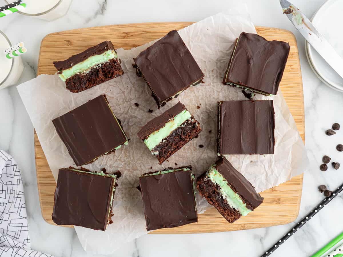 Nine Mint Chocolate Brownies with a green mint layer are arranged on parchment paper atop a wooden board, surrounded by a knife, chocolate chips, and glasses of milk on a marble surface.
