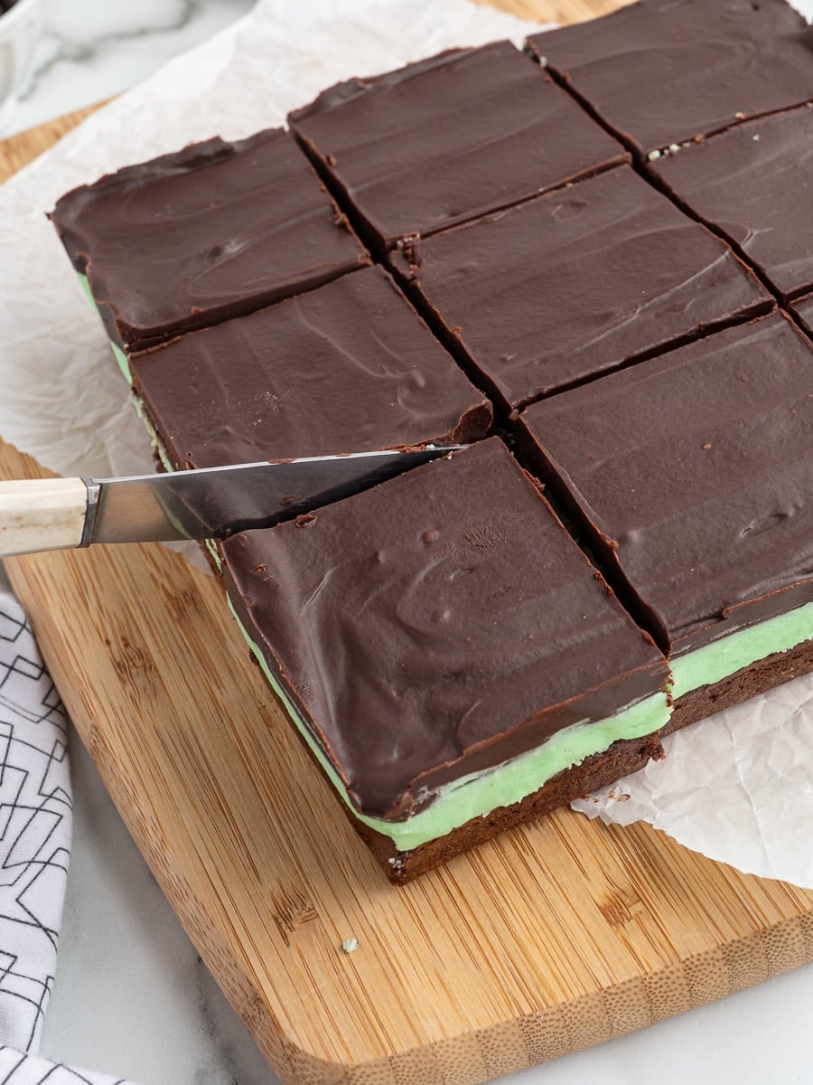 A knife slices into a square dessert with a chocolate top layer and a green mint layer, set on a wooden cutting board. These Mint Chocolate Brownies are cut into nine squares, with a white napkin nearby.