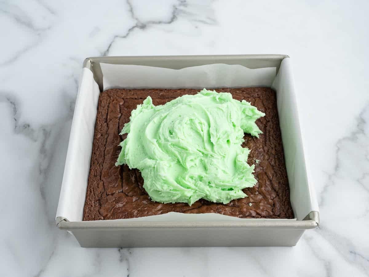 A square pan of Mint Chocolate Brownies sits on a marble surface, topped with a large mound of light green frosting in the center, ready to be spread. The pan is lined with parchment paper.