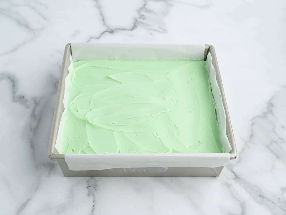 A square metal baking pan lined with parchment paper holds a smooth layer of light green batter, perfect for a Mint Chocolate Brownies Recipe, set on a white marble surface.