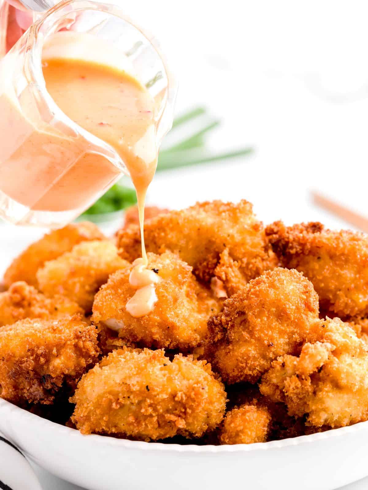 A hand pours creamy dipping sauce over a plate of golden, crispy Bang Bang Chicken nuggets.
