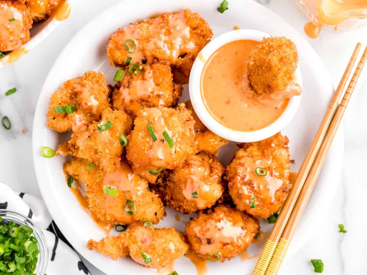 A plate of crispy fried shrimp topped with a tangy sauce and chopped green onions, served alongside a bowl of dipping sauce and a pair of wooden chopsticks on the side, drawing inspiration from the bold flavors found in the classic Bang Bang Chicken Recipe.