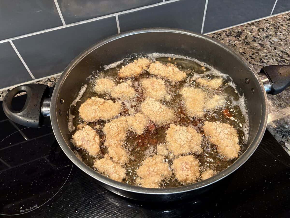 Several breaded chicken nuggets, reminiscent of a classic Bang Bang Chicken recipe, are being deep-fried in hot oil in a large black skillet on a stovetop, with bubbling oil surrounding the golden pieces.
