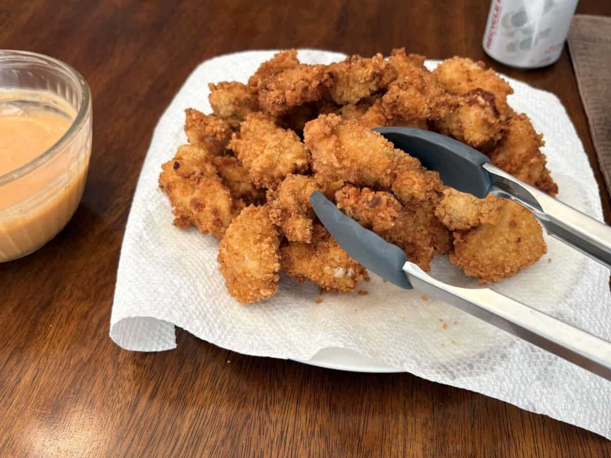 A plate of crispy, golden-brown Bang Bang Chicken nuggets rests on a paper towel, with metal tongs picking up a piece. A bowl of spicy orange dipping sauce sits on the side, bringing this Bang Bang Chicken recipe to life atop a wooden table.