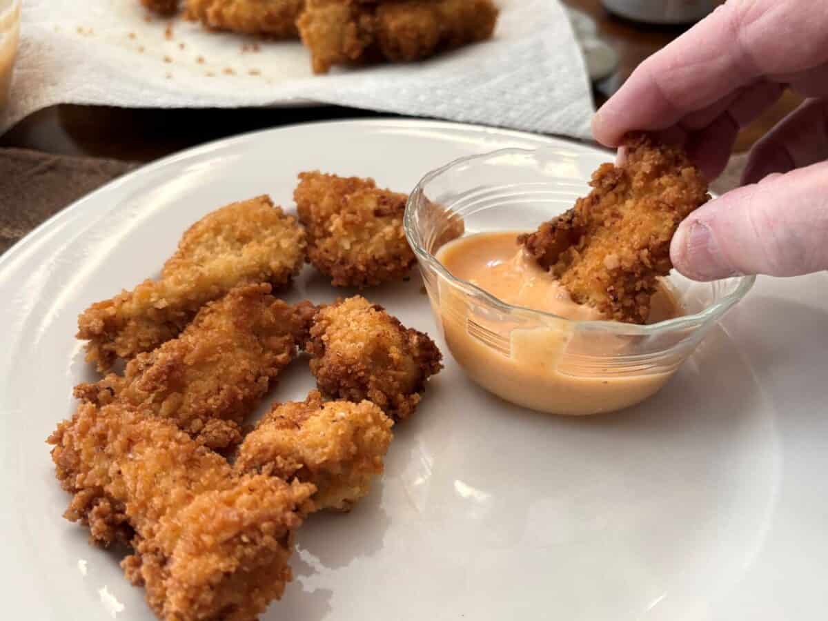 A hand dips a crispy breaded Bang Bang Chicken strip into a small glass bowl of orange dipping sauce, with more chicken strips on a white plate.