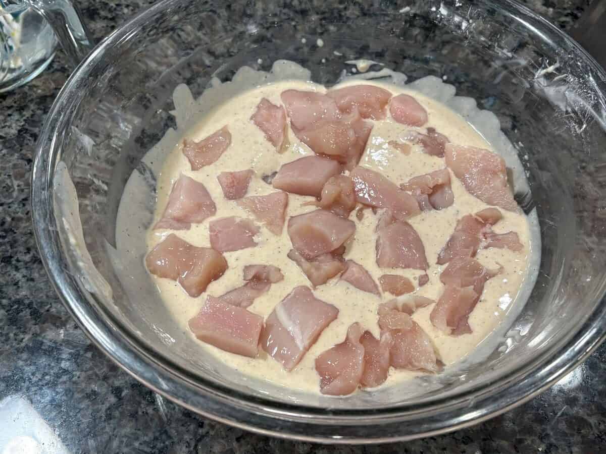 Raw chicken pieces are marinating in a creamy, seasoned mixture inside a clear glass bowl placed on a dark granite countertop, perfectly prepared for an authentic Bang Bang Chicken Recipe.
