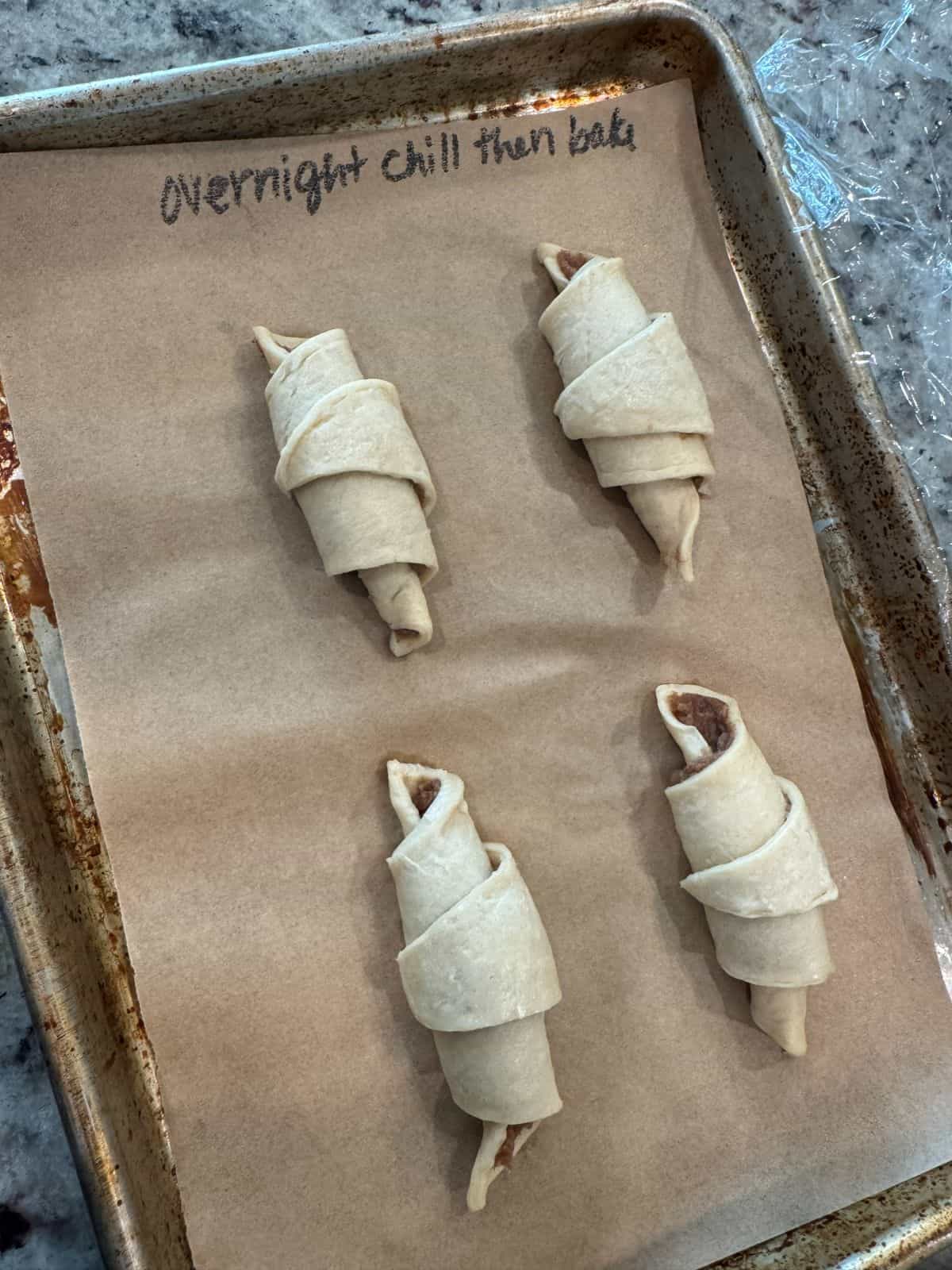 Four unbaked crescent rolls are arranged on a parchment-lined baking sheet, ready to become delicious Crescent Roll Cinnamon Rolls. “Overnight chill then bake” is handwritten at the top of the parchment paper, hinting at the simple yet irresistible Crescent Roll Cinnamon Rolls Recipe that awaits.