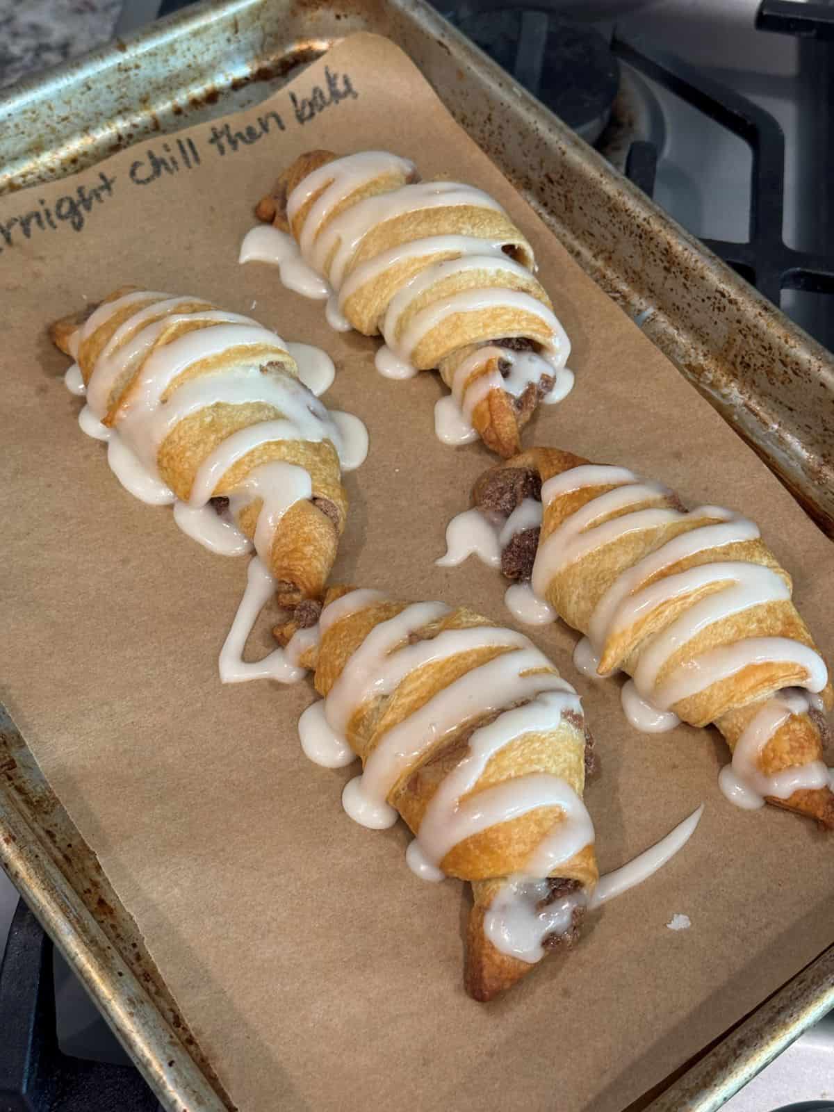 Four baked crescent roll cinnamon rolls topped with white glaze on a parchment-lined baking sheet beside a stovetop.