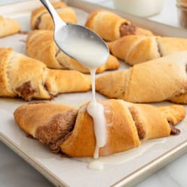 A spoon drizzles white icing onto a freshly baked Crescent Roll Cinnamon Roll on a parchment-lined baking sheet, with more rolls in the background, showcasing the irresistible results of this easy Crescent Roll Cinnamon Rolls Recipe.