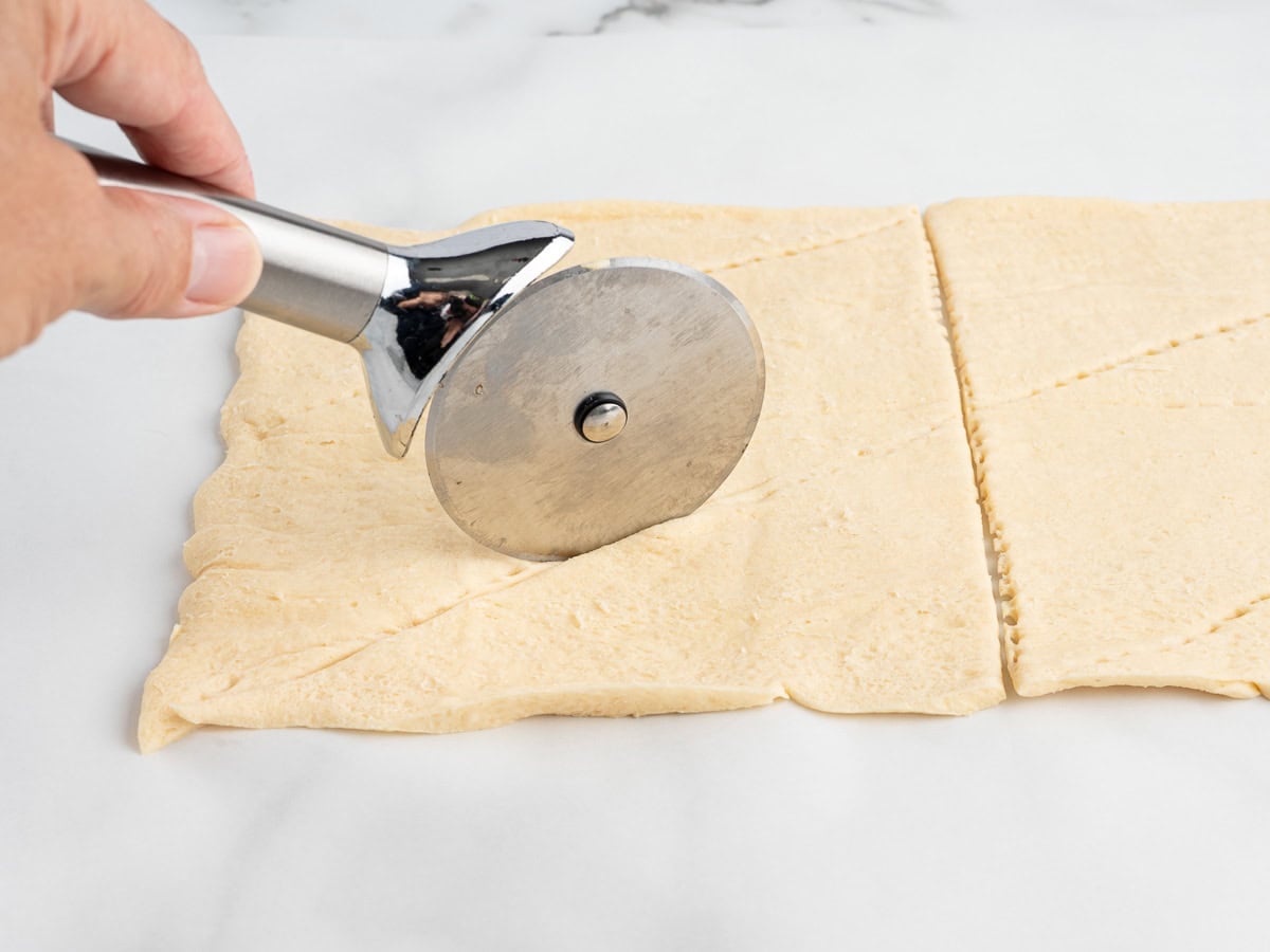 A hand runs a pizza cutter along the perforations in a sheet of crescent roll dough on a white work surface.