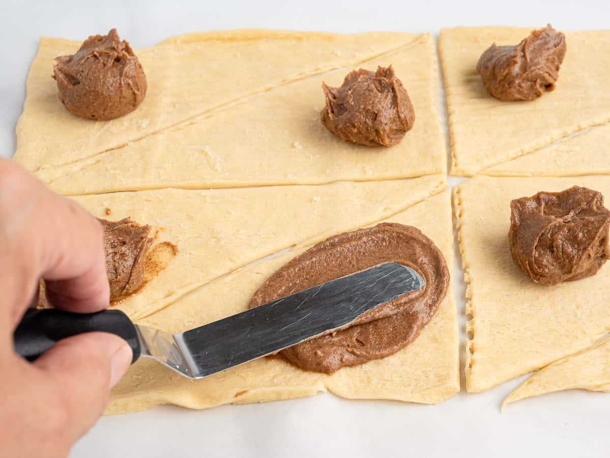 A hand spreads the cinnamon and brown sugar filling onto a triangle of crescent roll dough with an offset spatula, with more filled triangles beside it.