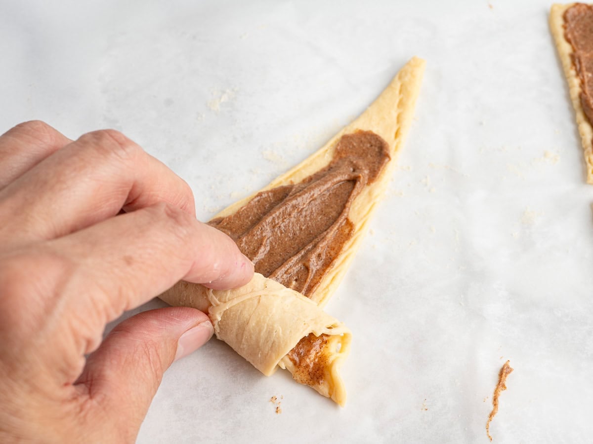 A hand rolls up a triangle of dough spread with a brown cinnamon mixture on parchment paper, preparing a delectable Crescent Roll Cinnamon Rolls pastry.