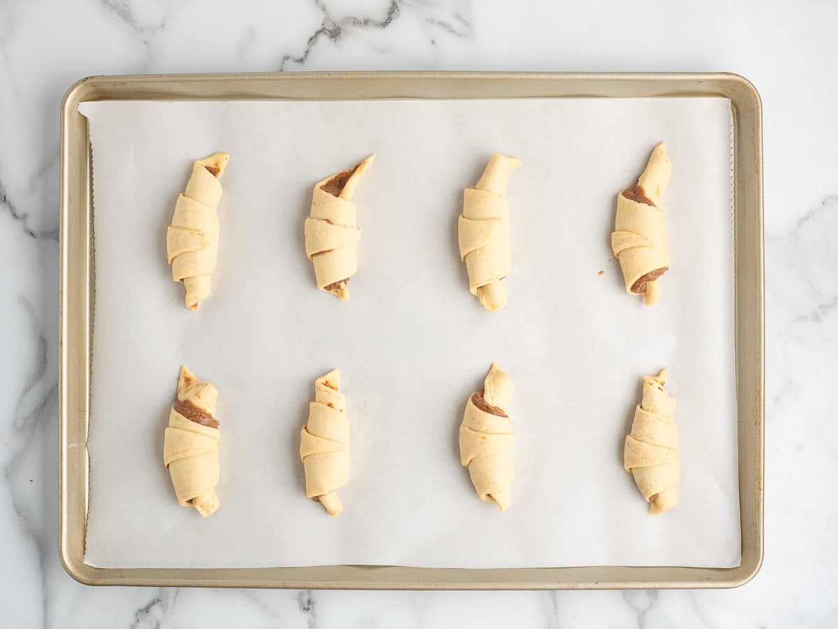 A baking sheet lined with parchment paper holds eight unbaked Crescent Roll Cinnamon Rolls, evenly spaced on a marble countertop, ready to follow your favorite Crescent Roll Cinnamon Rolls Recipe.