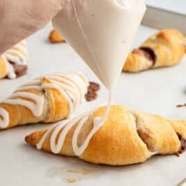 A hand pipes white icing onto a freshly baked crescent roll on a parchment-lined baking sheet, with more Crescent Roll Cinnamon Rolls visible in the background—perfectly showcasing this easy and delicious Crescent Roll Cinnamon Rolls Recipe.