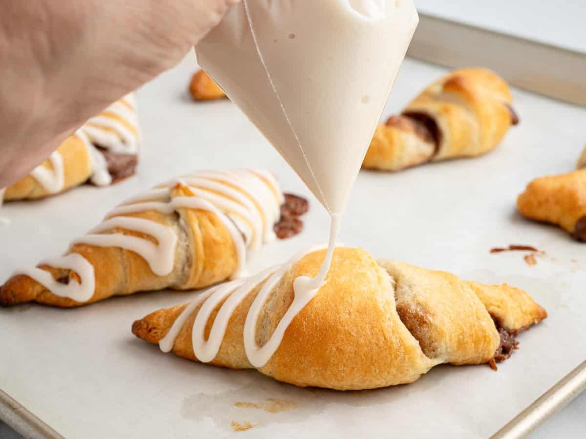 A hand pipes white glaze over a baked crescent roll cinnamon roll on a parchment-lined baking sheet, with more rolls behind it.