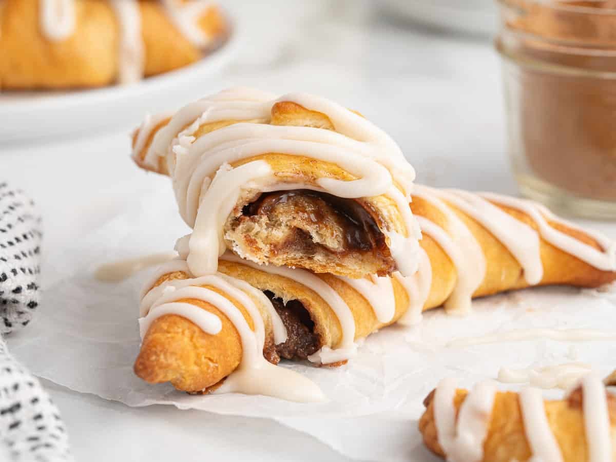 Two crescent roll cinnamon rolls drizzled with white icing on white parchment paper, one broken open to show the cinnamon and brown sugar filling inside, with a striped towel and a glass jar behind them.