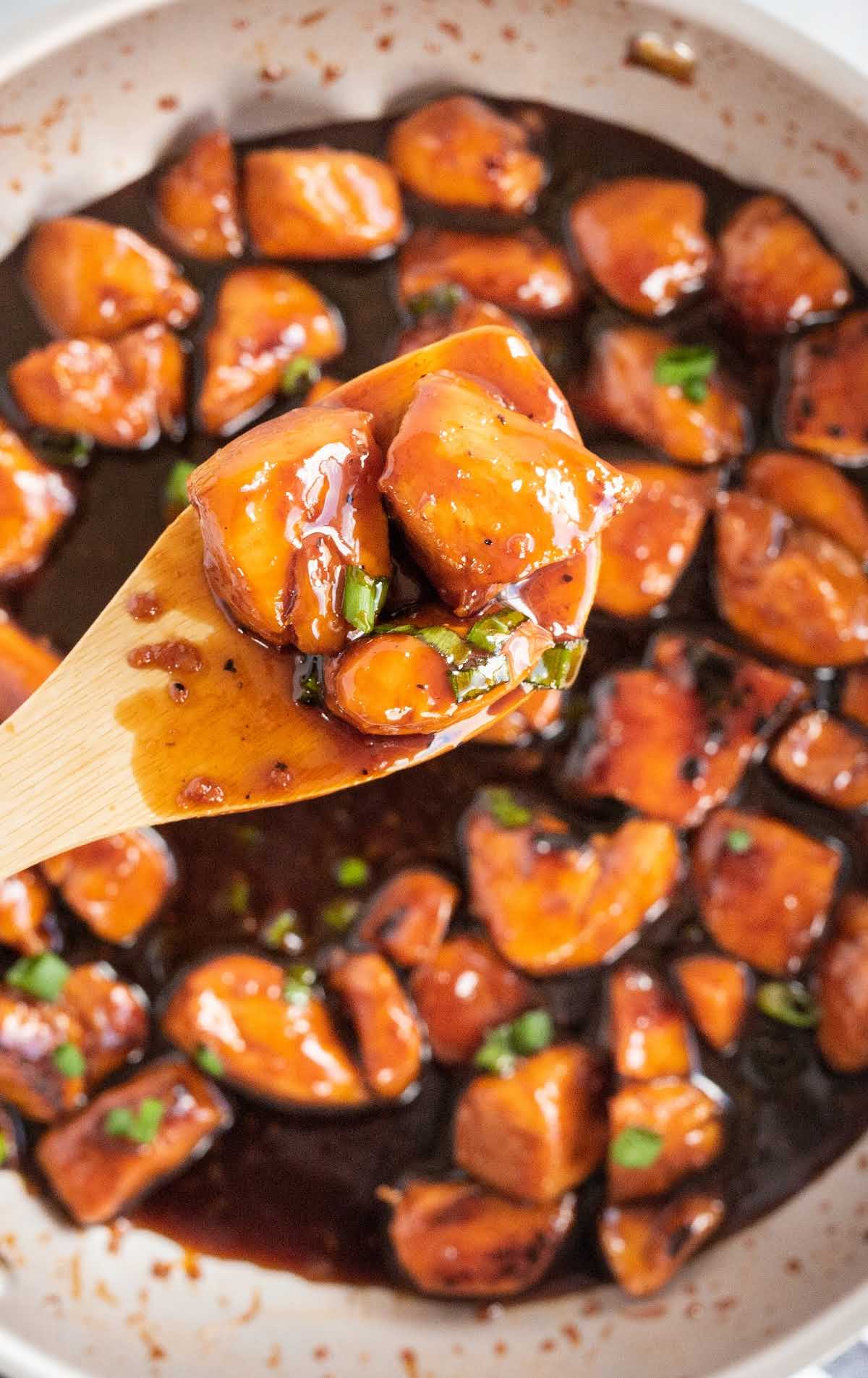 close up overhead shot of bourbon chicken topped with diced green onions in a skillet with a wooden spoon