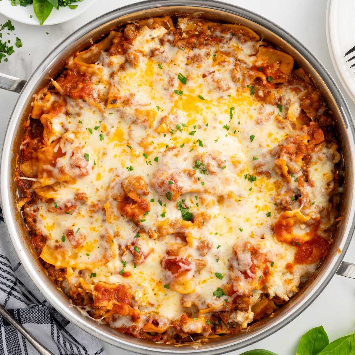 Overhead view of cooked skillet lasagna in a metal pan, topped with melted cheese and fresh herbs, placed on a marble surface.