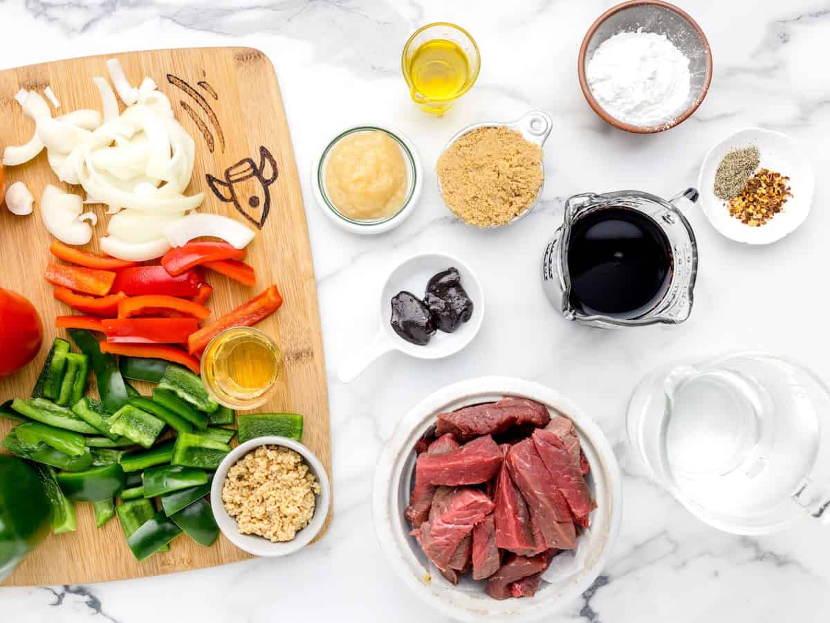 Sliced vegetables, raw beef strips, and various sauces and seasonings are arranged on a white surface with a wooden cutting board, ready for cooking a delicious Slow Cooker Pepper Steak.