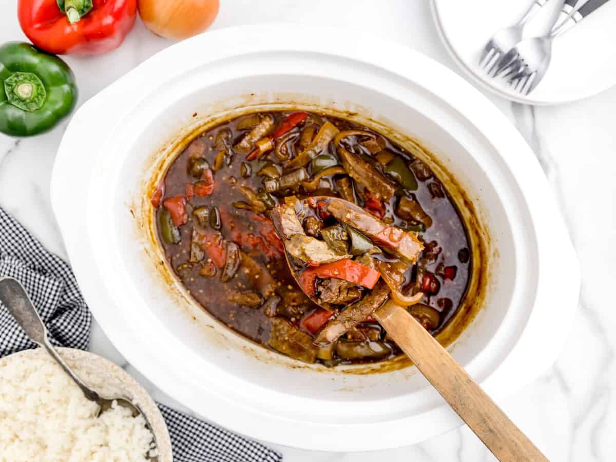 A white slow cooker filled with Slow Cooker Pepper Steak—tender beef and bell peppers simmered in a flavorful sauce—is shown with a wooden spoon lifting a hearty portion. Rice, utensils, a green pepper, and a red pepper are arranged nearby on the marble surface.
