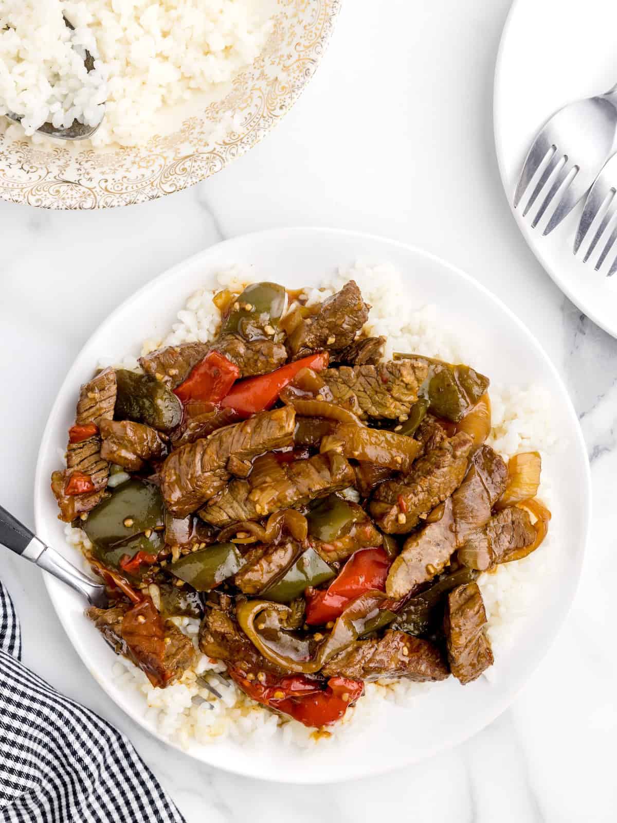 A plate of white rice topped with tender Slow Cooker Pepper Steak, featuring stir-fried beef, red and green bell peppers, and onions, with a fork on the side. A bowl of rice and utensils are nearby on a marble surface.