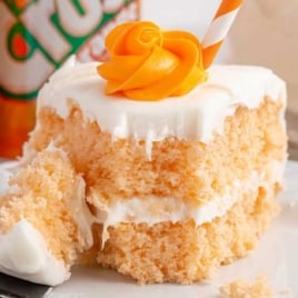 Fork taking a bite of fluffy orange soda cake layered with white frosting, with cans of orange Crush in the background.