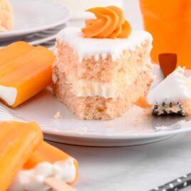 a slice of cake topped with frosting on a plate surrounded by orange ice cream popsicles