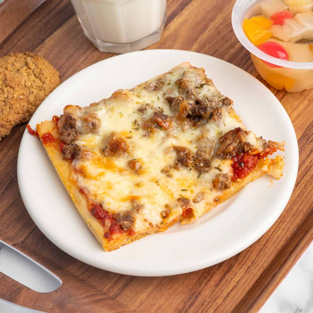 A slice of sausage pizza, reminiscent of classic Rectangle School Pizza, sits on a white plate alongside a cookie, a cup of fruit, and a glass of milk on a wooden tray.