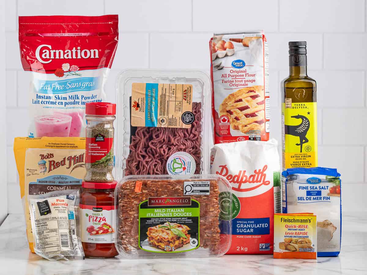 A variety of grocery items essential for making a delicious Rectangle School Pizza, including ground beef, sugar, olive oil, flour, milk powder, Italian sausage, yeast, salt, spices, canned tomatoes, and pizza sauce, are displayed on a white kitchen counter.