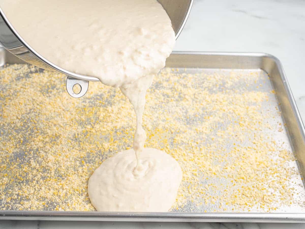 Batter is being poured from a mixing bowl onto a baking sheet sprinkled with cornmeal, on a marble countertopโjust like the classic Rectangle School Pizza Recipe.