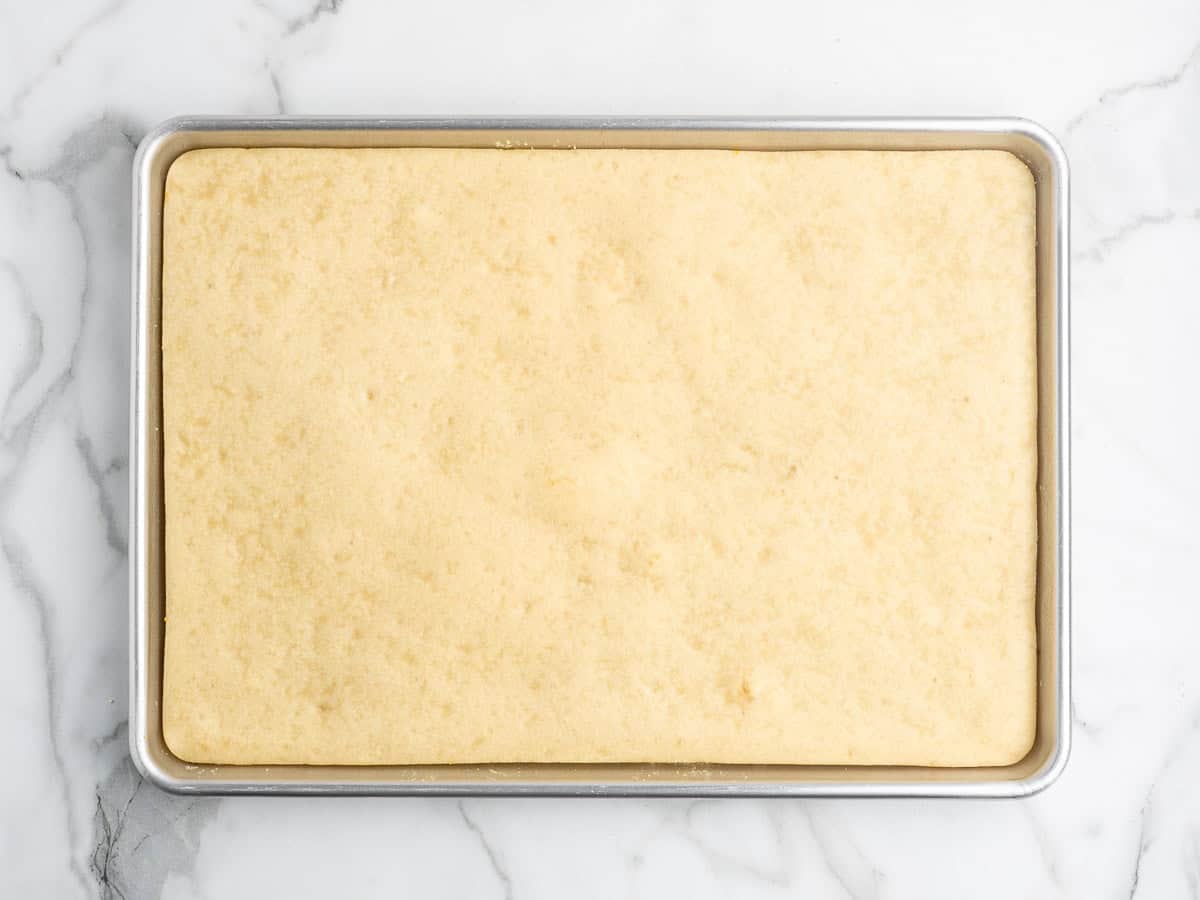 A rectangular baking pan filled with an unbaked, pale yellow dough sits on a white marble surface, ready to become a classic Rectangle School Pizza.