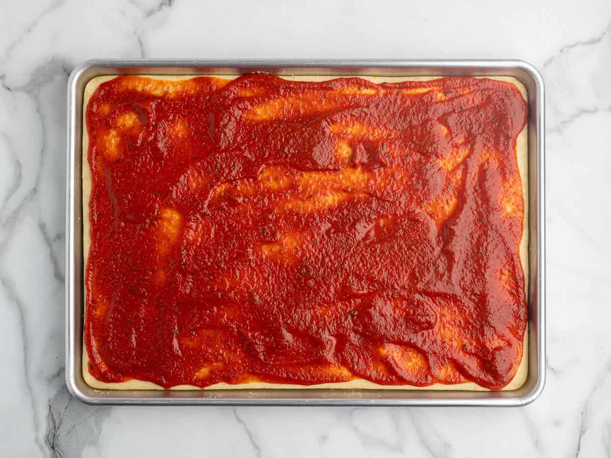A rectangular baking sheet with pizza dough spread with tomato sauce, ready to become a classic Rectangle School Pizza on a marble surface.